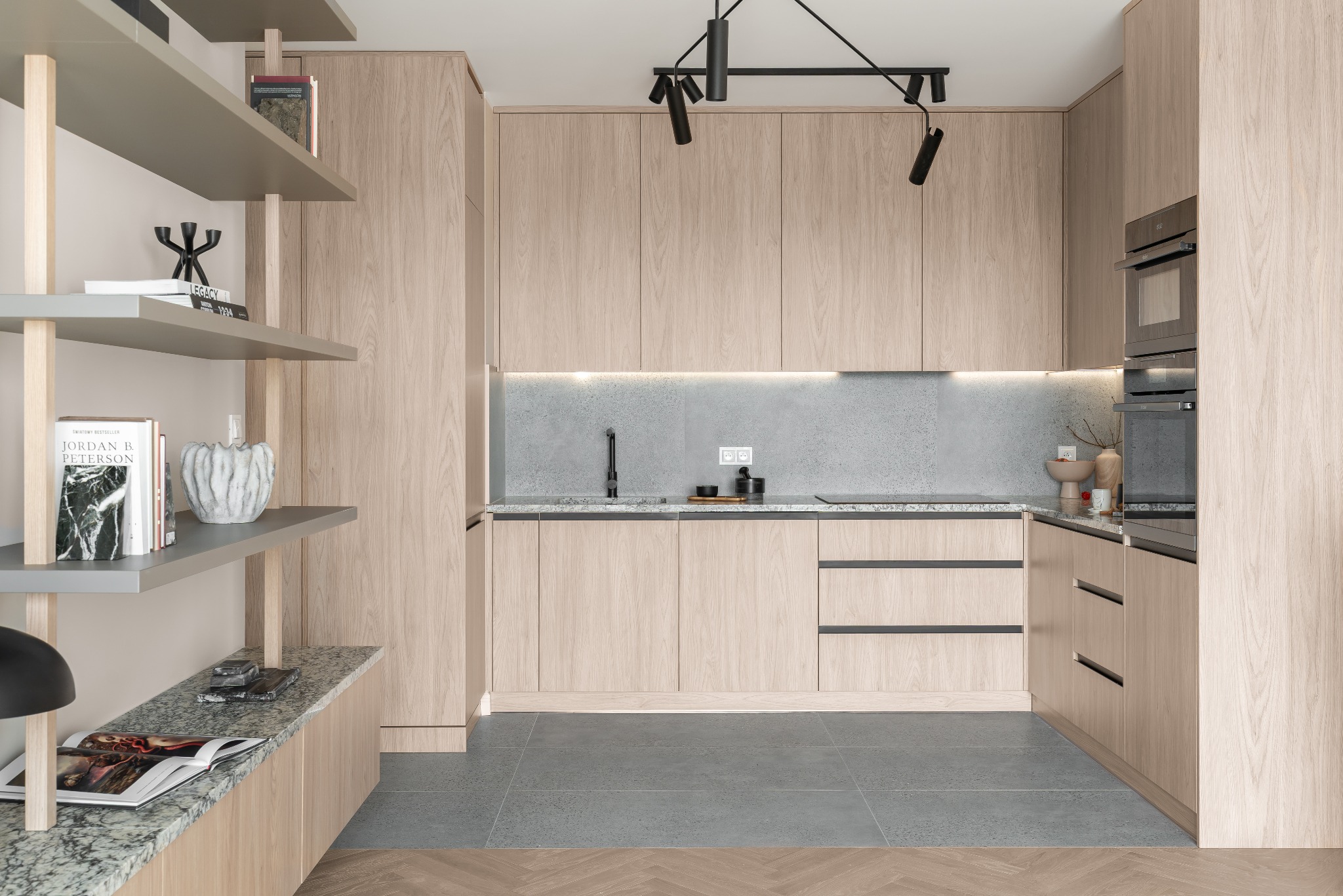 Jasna, nowoczesna kuchnia z drewnianymi szafkami, szarym blatem i płytkami. Oświetlenie punktowe. Na półkach książki i dekoracje. W tle sprzęt AGD w zabudowie.