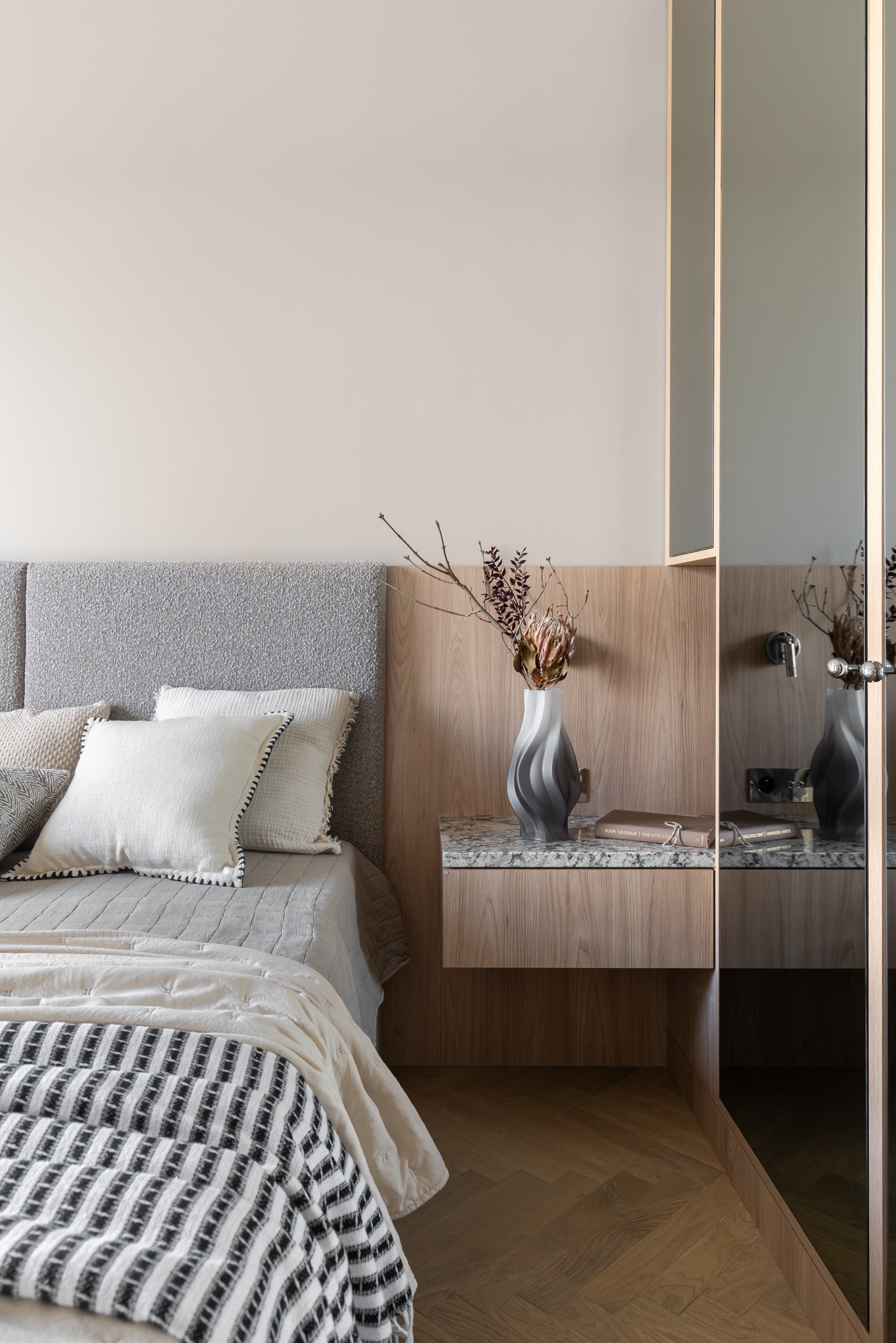 Nowoczesna sypialnia: łóżko z szarym wezgłowiem, stolik nocny z marmurowym blatem i wazonem z suszem, parkiet w jodełkę, lustrzane drzwi szafy.