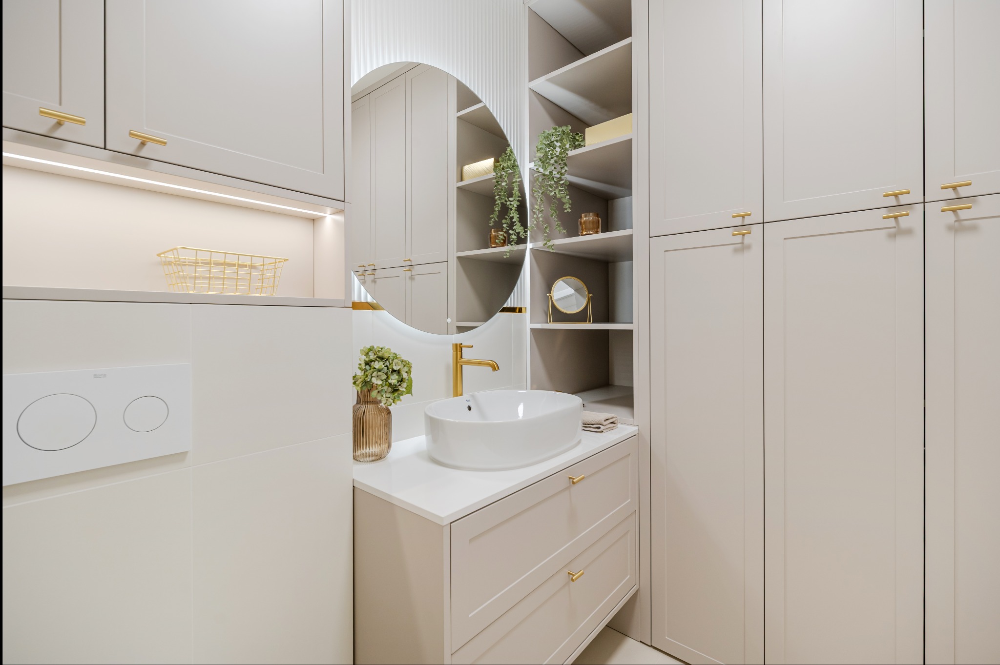 Jasna łazienka z owalnym lustrem z podświetleniem, białą umywalką nablatową i złotą armaturą. Zabudowa meblowa z szafkami i półkami.