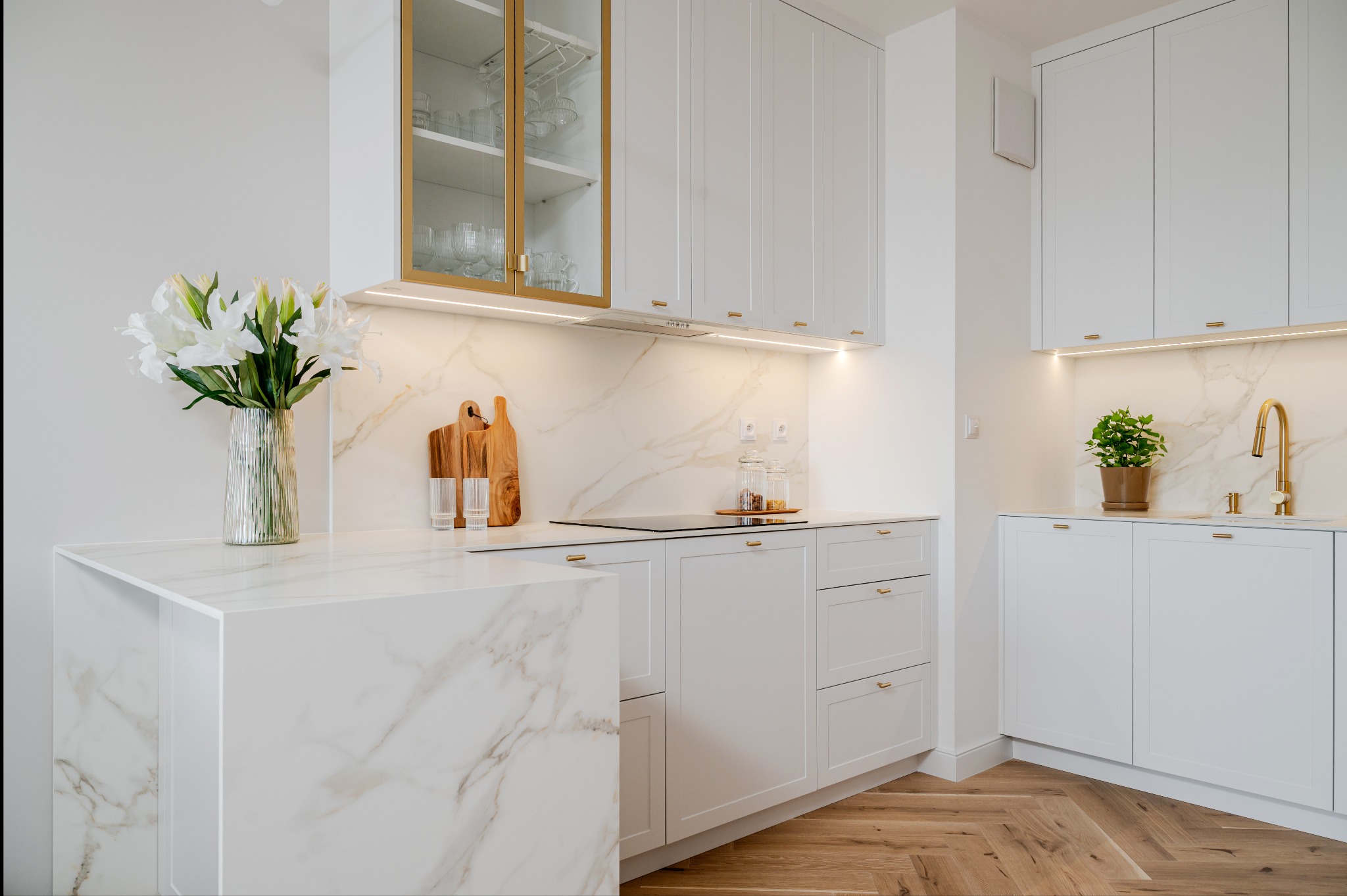 Elegancka, jasna kuchnia z marmurowym blatem i drewnianą podłogą w jodełkę. Białe szafki z delikatnymi złotymi uchwytami. Wazon z liliami na wyspie.