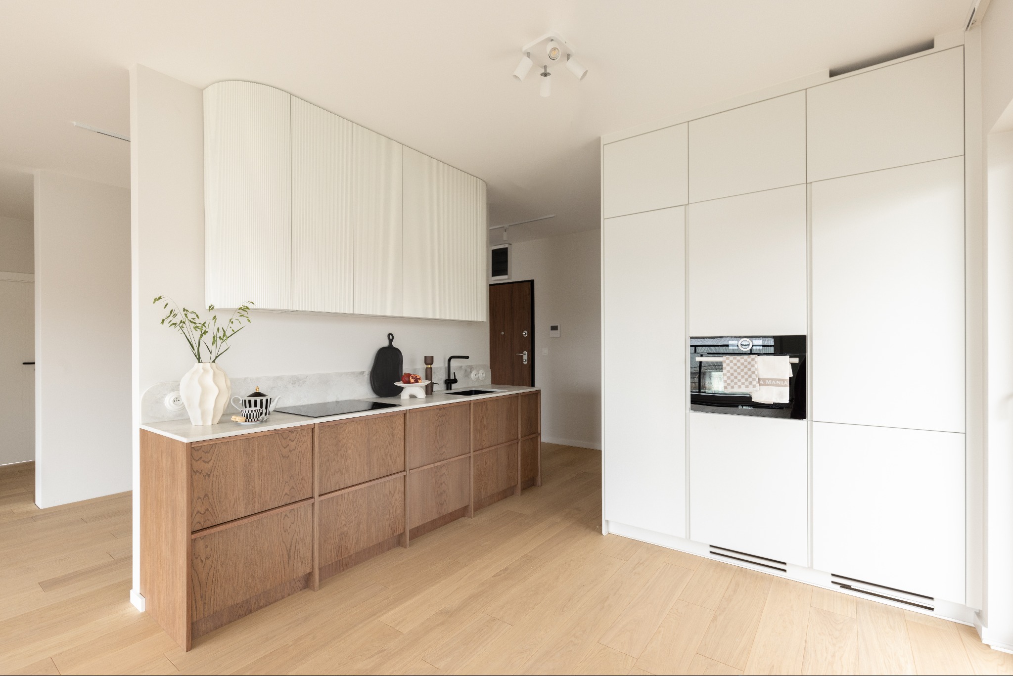 Nowoczesna, jasna kuchnia z drewnianymi szafkami i białymi górnymi szafkami z ryflowanym frontem. Na blacie dekoracje i czajnik. Parkiet na podłodze.