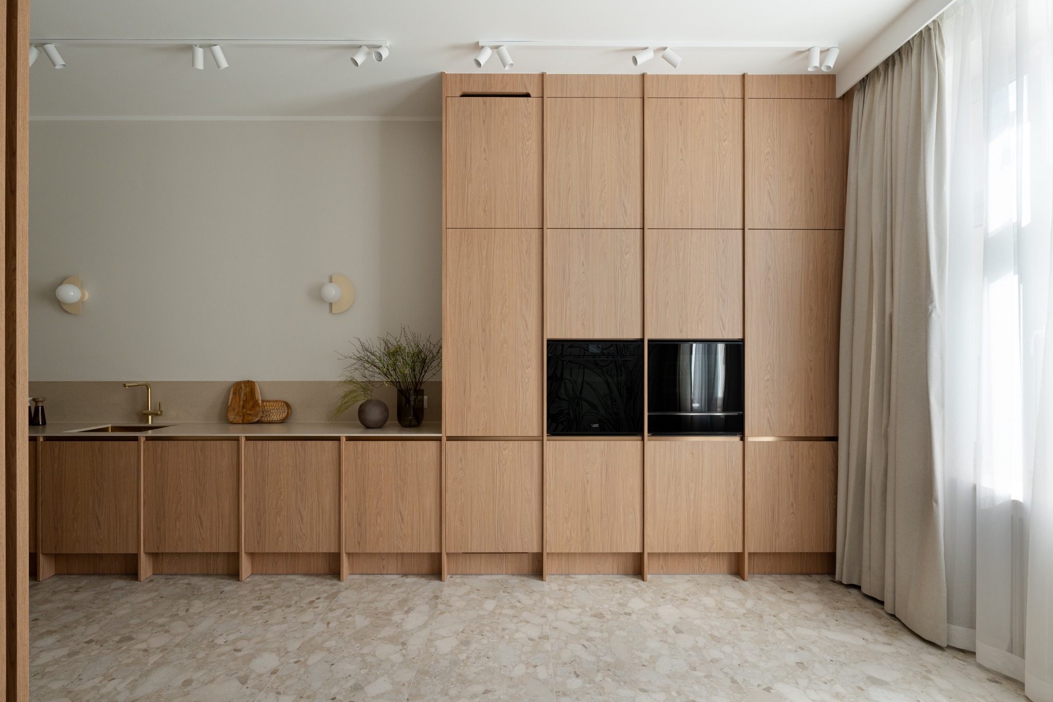 Nowoczesna zabudowa kuchenna z drewnianymi frontami, wbudowanym piekarnikiem i płytą. Beżowe ściany, marmurowa podłoga i subtelne oświetlenie tworzą spójną całość.