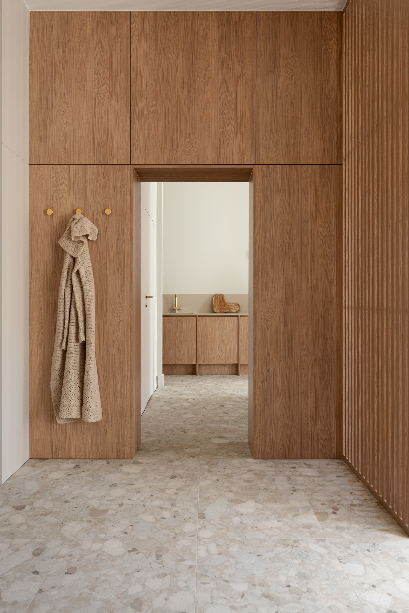 Nowoczesny hol z drewnianą zabudową szaf, dekoracyjną ścianką z listew i podłogą z płytek lastryko. W tle widoczna łazienka.