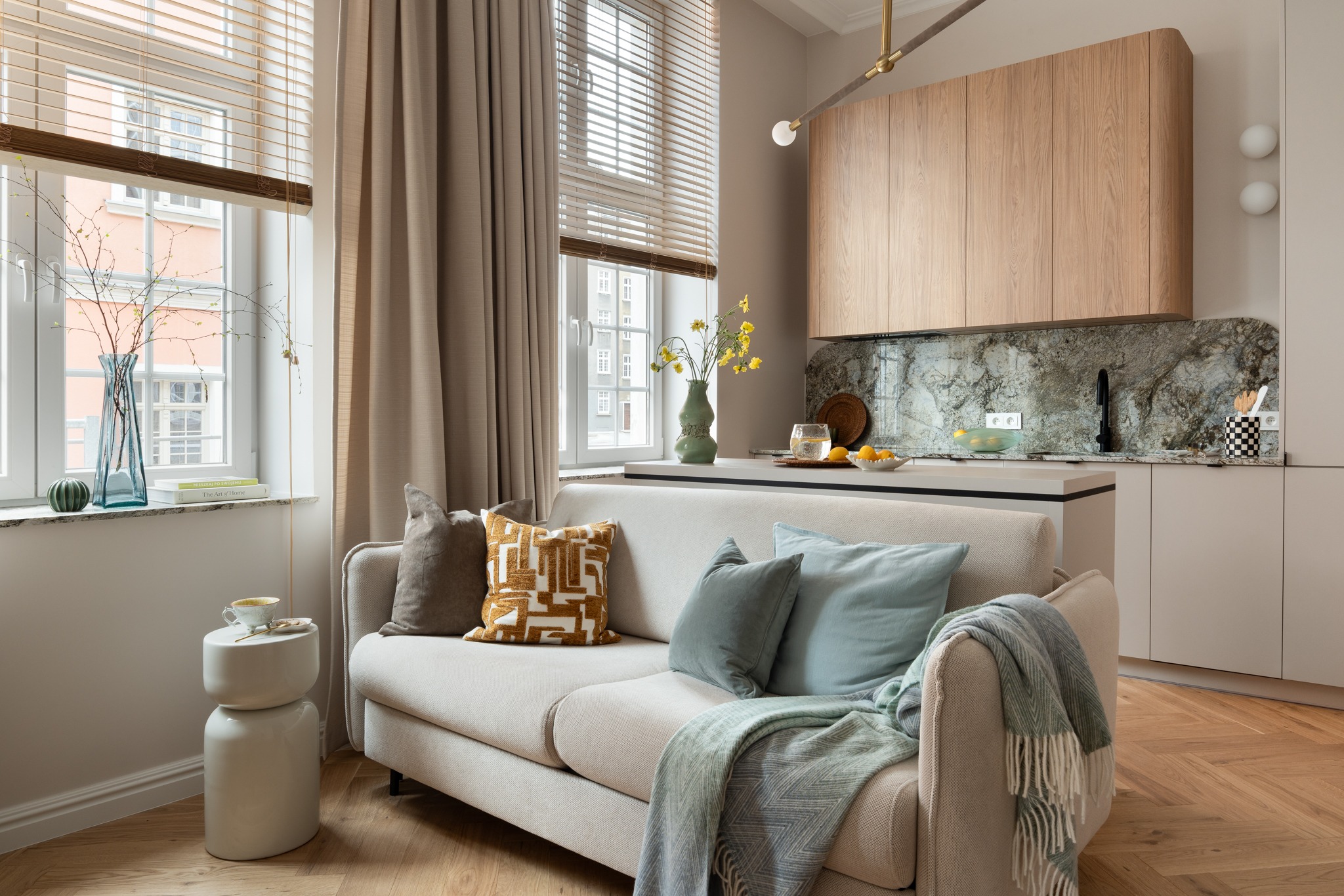 Jasne wnętrze: sofa z poduszkami i pledem, okno z żaluzjami i zasłonami, drewniana podłoga w jodełkę, marmurowy blat kuchenny. Wazon z kwiatami na blacie.