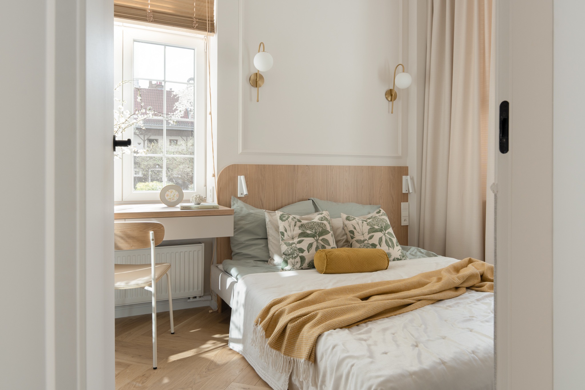 Jasna sypialnia z drewnianym zagłówkiem, biurkiem przy oknie i parkietem. Wnętrze w neutralnych barwach, oświetlone lampami ściennymi i naturalnym światłem.