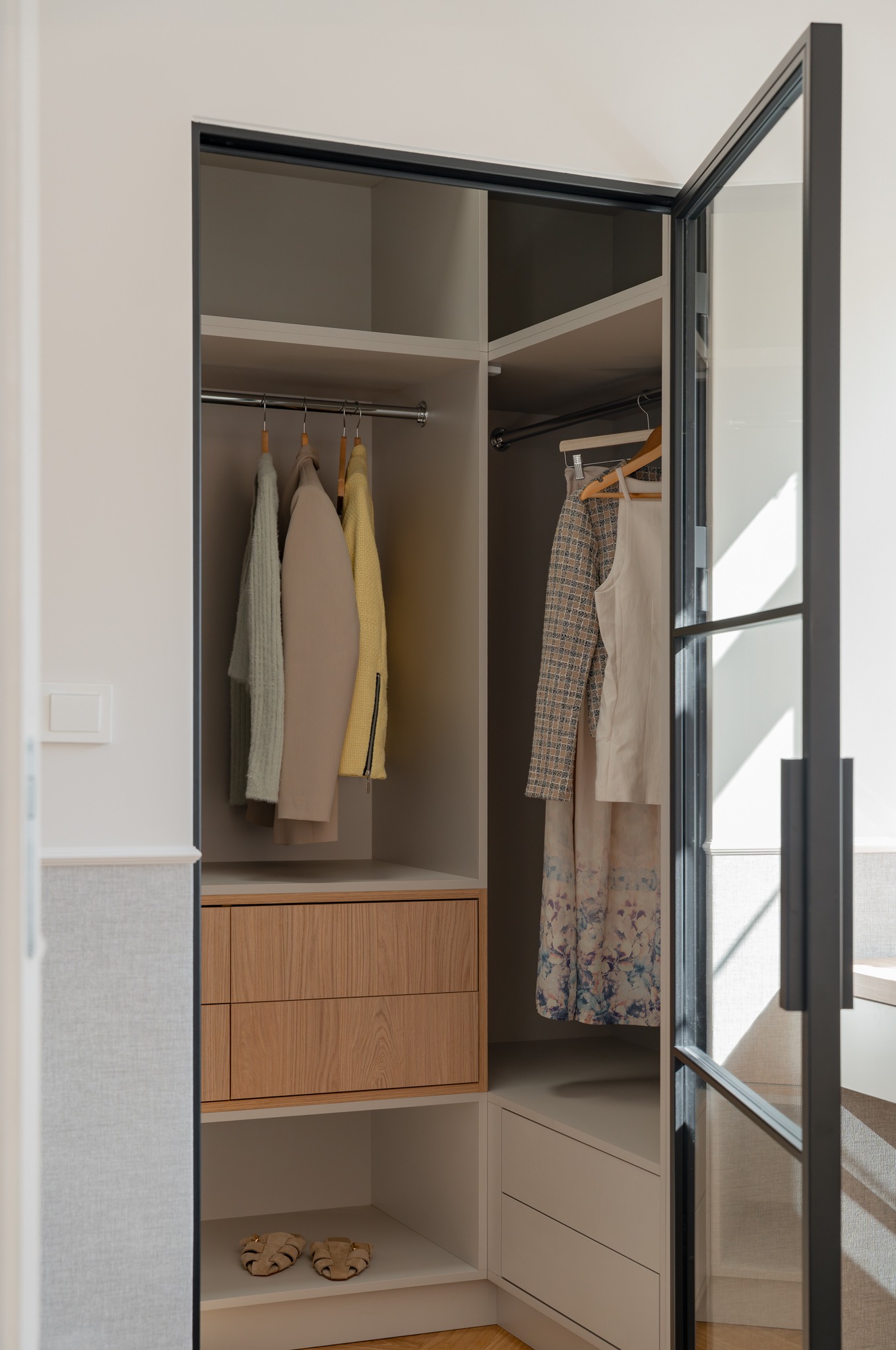 Nowoczesna garderoba narożna z przeszklonymi drzwiami w czarnej ramie. Ubrania wiszą na wieszakach, widoczne drewniane szuflady i półki. Jasne, naturalne światło.