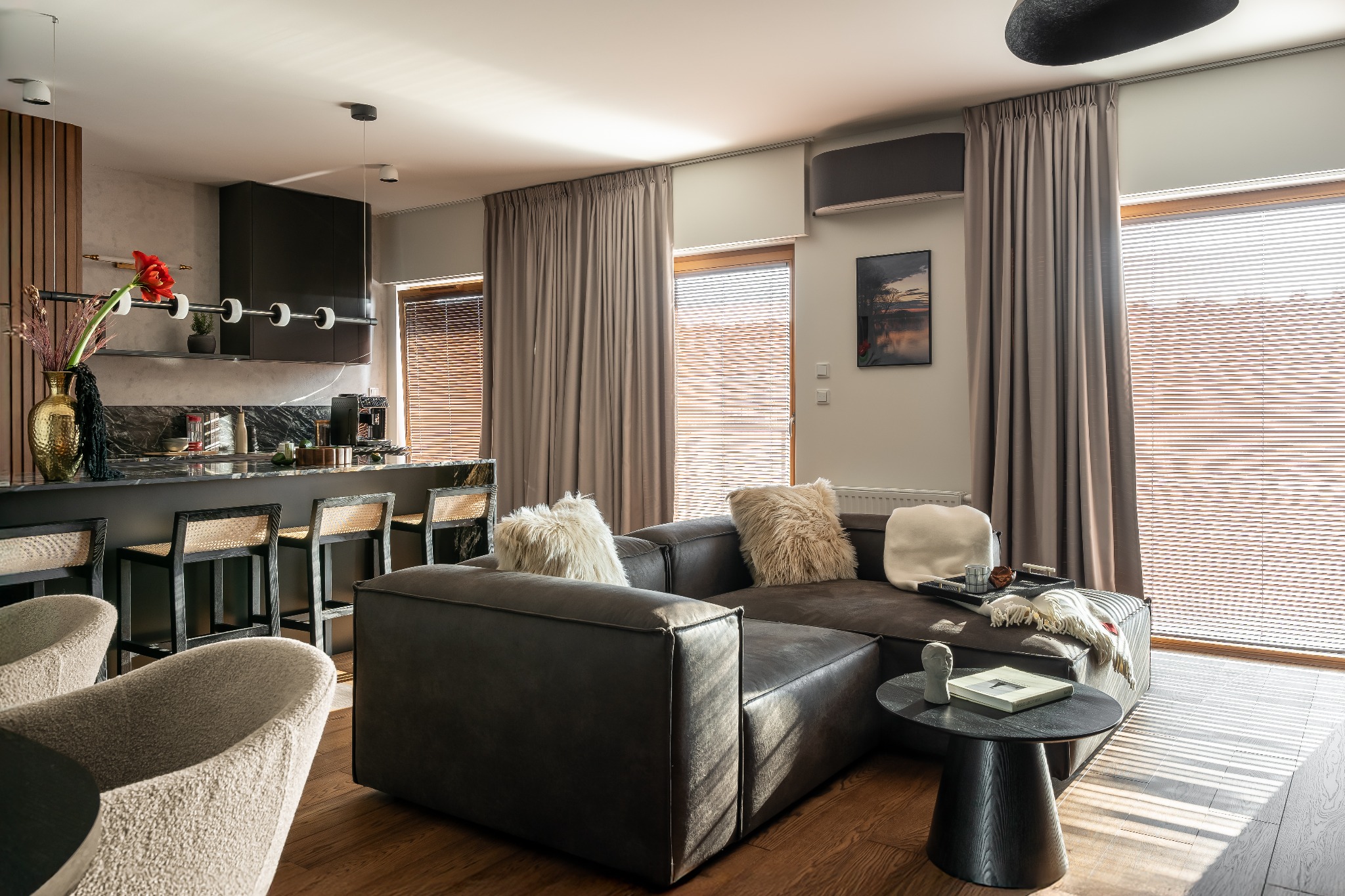 Stylowe wnętrze: ciemna kuchnia z wyspą i hokerami, szara sofa z poduszkami, drewniana podłoga, żaluzje w oknach. Nowoczesny wystrój.