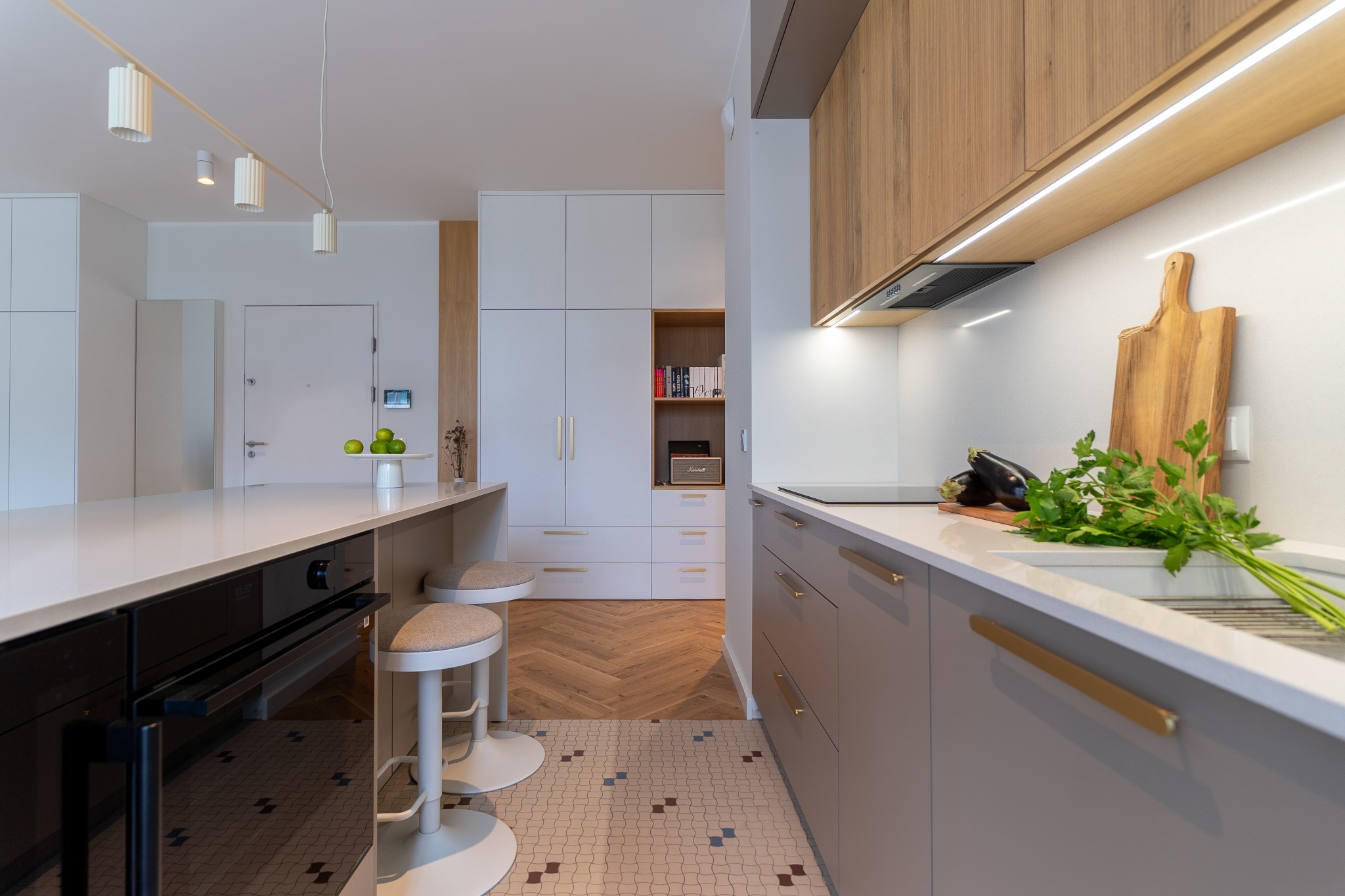 Nowoczesna kuchnia z wyspą, szare szafki z złotymi uchwytami, drewniane akcenty, oświetlenie LED, parkiet w jodełkę i dekoracyjna podłoga. Minimalistyczny design.