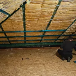 Pracownik w czarnym ubraniu klęczy na podłodze poddasza, montując izolację termiczną z wełny mineralnej pomiędzy zielonymi belkami konstrukcyjnymi dachu za pomocą wkrętarki. Na podłodze leży młotek...