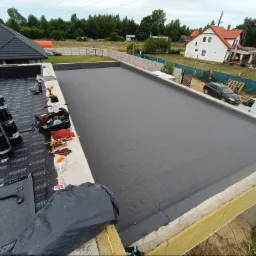 Widok z góry na płaski dach pokryty nową papą termozgrzewalną. Obok leżą rolki papy Izobud, narzędzia dekarskie i torba narzędziowa. W tle zabudowa jednorodzinna i zieleń.