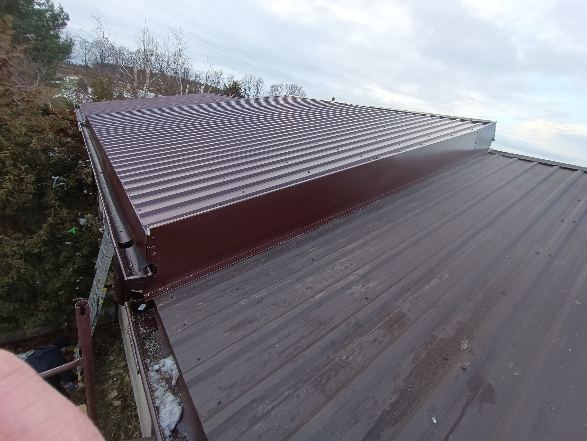 Nowy, brązowy dach z blachy trapezowej z widoczną obróbką blacharską, zamontowany na budynku z widocznymi drzewami w tle.