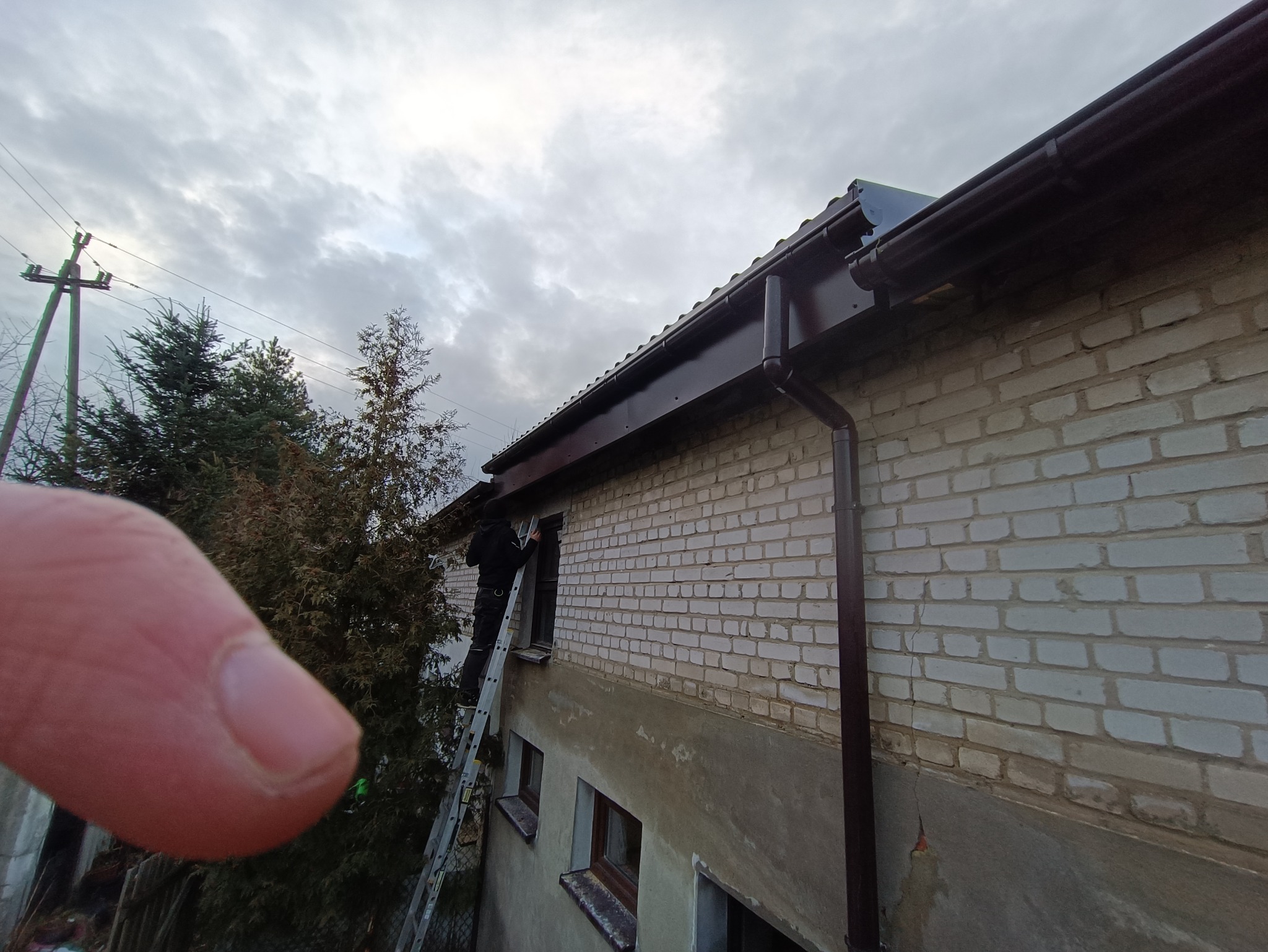 Pracownik na drabinie przy budynku z białej cegły, montaż lub naprawa rynny w kolorze ciemnego brązu, widoczny fragment palca w kadrze, pochmurne niebo w tle.