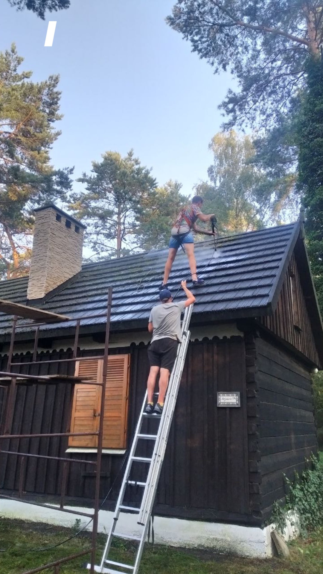 Dwóch mężczyzn czyści dach drewnianego domu za pomocą myjki ciśnieniowej, jeden na dachu, drugi na drabinie, w tle drzewa.