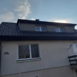 Dom z ciemnoszarą blachodachówką, numer 39A na elewacji i widocznym tęczowym halo wokół słońca na niebie.