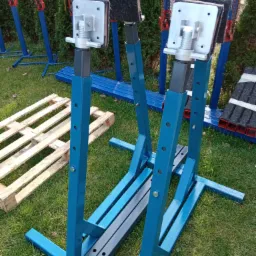 Dwa niebieskie stojaki ślusarskie z regulacją wysokości na trawie, obok drewniana paleta i zwinięte metalowe profile.
