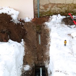 ELEKTRON Usługi Elektryczne Oskar Zaremba - Wykop w ziemi, częściowo pokryty śniegiem, z uziomem w kształcie litery J w głębi, widoczna zielona żyła uziemiająca przy ścianie budynku, śrubokręt na śniegu i fragment muru z cegły.