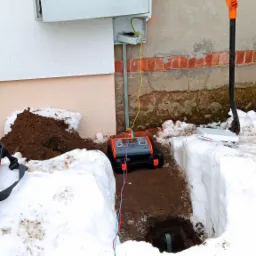 Pomiar rezystancji uziomu przy skrzynce elektrycznej na zewnątrz budynku w zimowych warunkach, widoczny miernik, wykop i łopata.