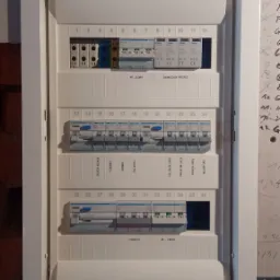 Rozdzielnica elektryczna marki Hager, zamontowana w białej obudowie w ścianie z cegły i płyty gipsowej, widoczne zabezpieczenia i opisy obwodów.