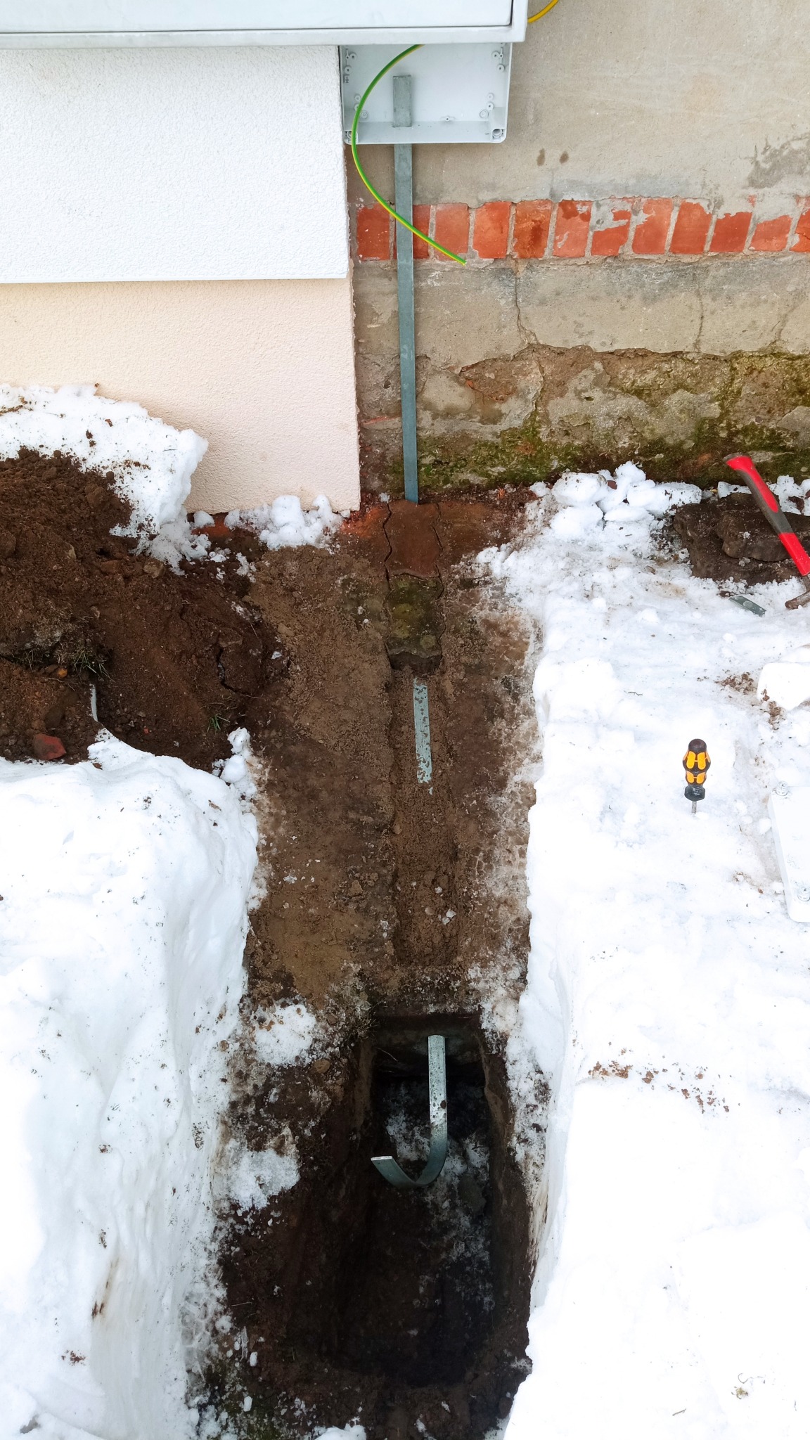 Wykop w ziemi, częściowo pokryty śniegiem, z uziomem w kształcie litery J w głębi, widoczna zielona żyła uziemiająca przy ścianie budynku, śrubokręt na śniegu i fragment muru z cegły.