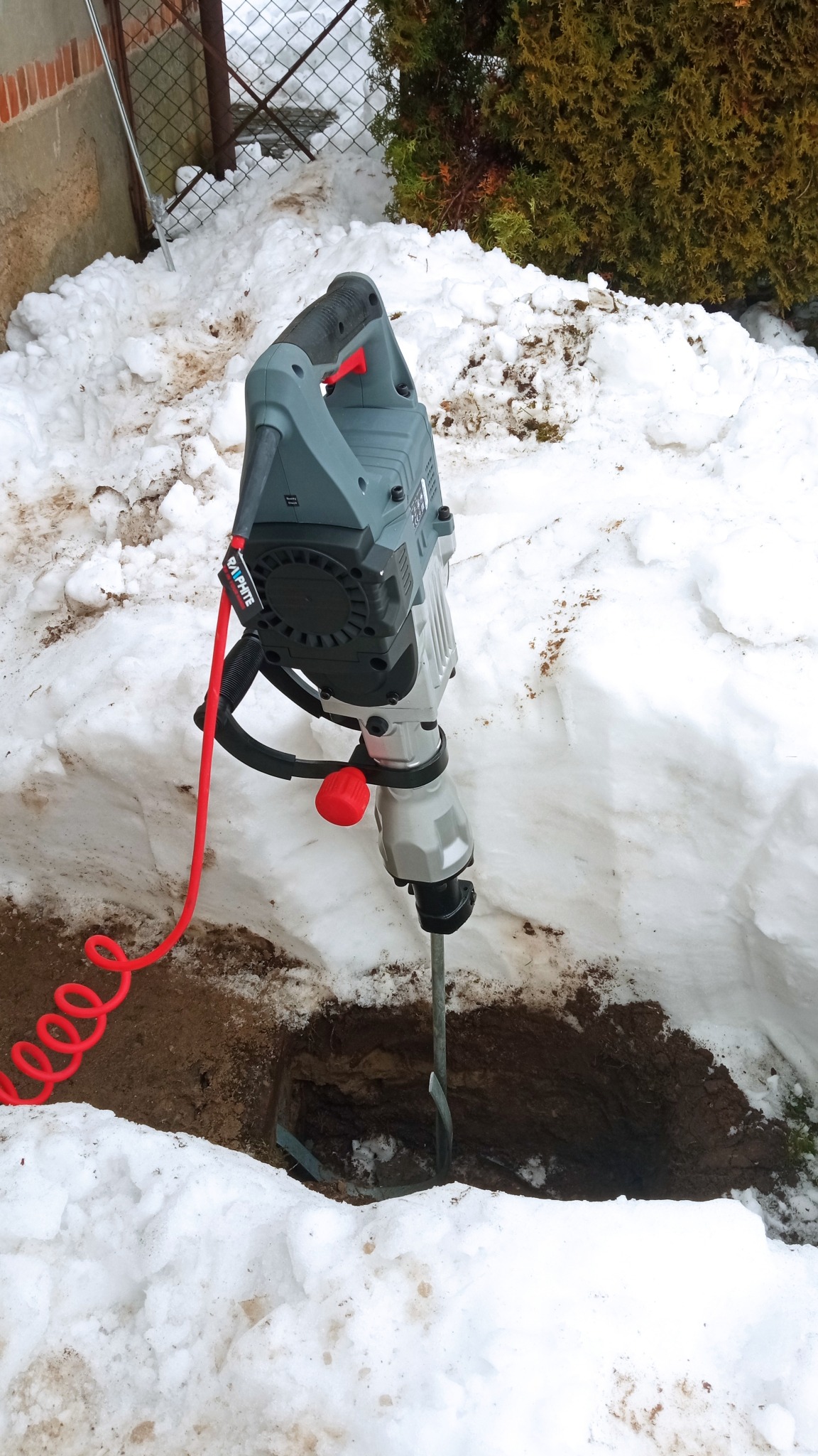 Wiertnica udarowa Graphite wbita w ziemię, otoczona śniegiem, czerwony kabel zasilający zwinięty w spiralę, widoczne ślady wykopu.