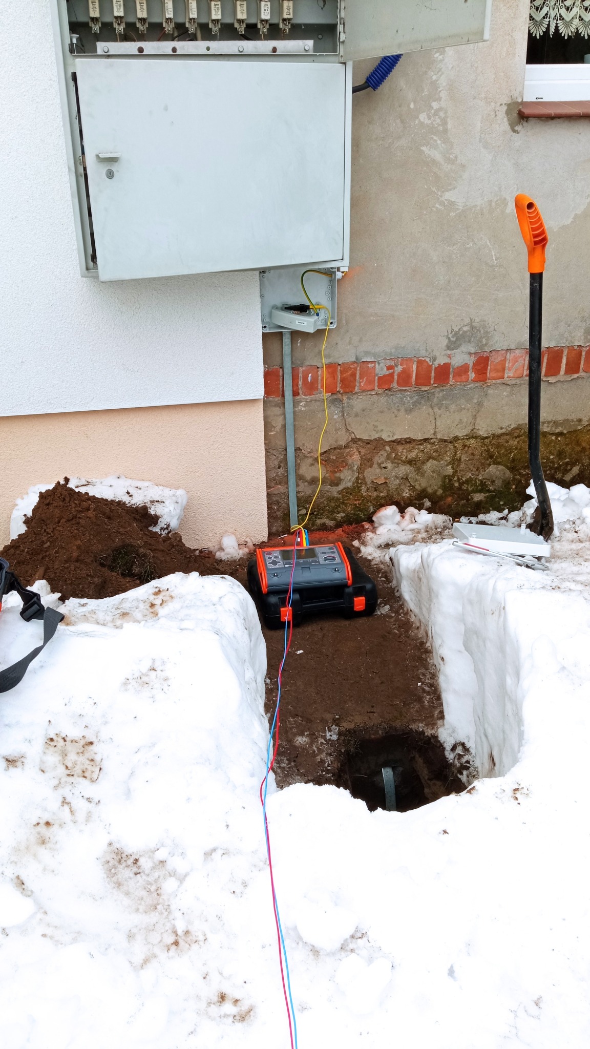 Pomiar rezystancji uziomu przy skrzynce elektrycznej na zewnątrz budynku w zimowych warunkach, widoczny miernik, wykop i łopata.