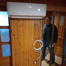Kamil-Instalacje KAMIL SIKORA - Biały klimatyzator Rotenso zamontowany na drewnianej ścianie z widocznym kablem zasilającym i fragmentem sylwetki mężczyzny odbijającej się w lustrze drzwi przesuwnych