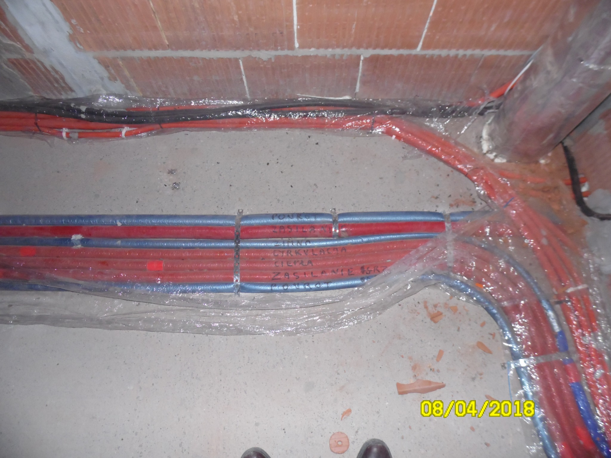 Instalacja rur grzewczych pod podłogą, zabezpieczonych folią, z widocznymi napisami na rurach oraz fragmentem muru z cegły i butami w dolnej części kadru.