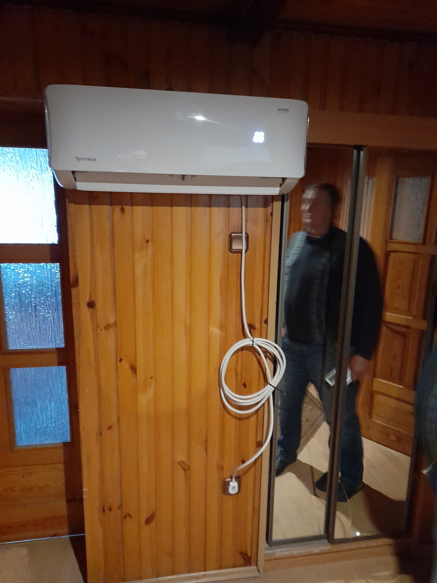 Biały klimatyzator Rotenso zamontowany na drewnianej ścianie z widocznym kablem zasilającym i fragmentem sylwetki mężczyzny odbijającej się w lustrze drzwi przesuwnych