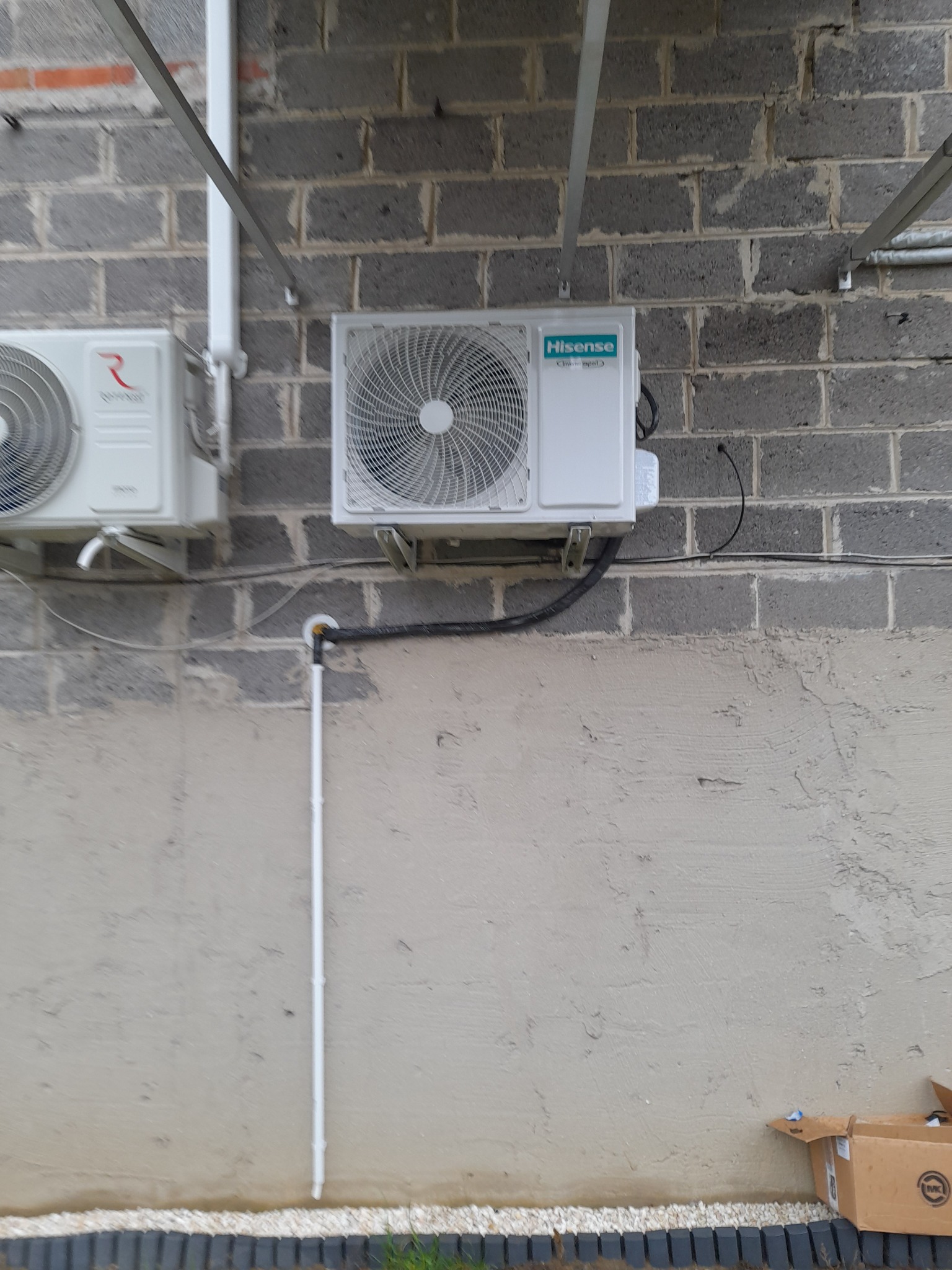 Dwa klimatyzatory zamontowane na szarej ścianie z bloczków betonowych, widoczne rury odprowadzające skropliny i przewody elektryczne.