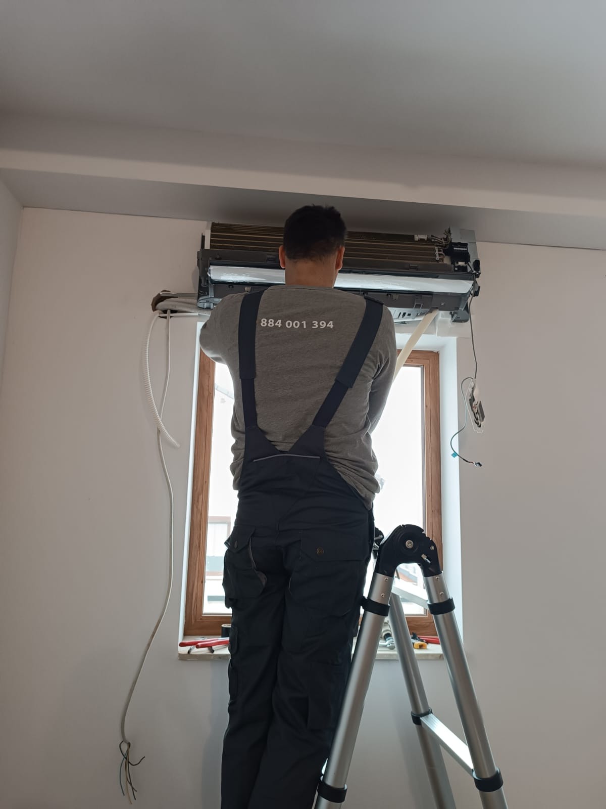 Instalator na drabinie montuje klimatyzator ścienny, widoczne przewody, okno w tle, numer telefonu na koszulce instalatora.
