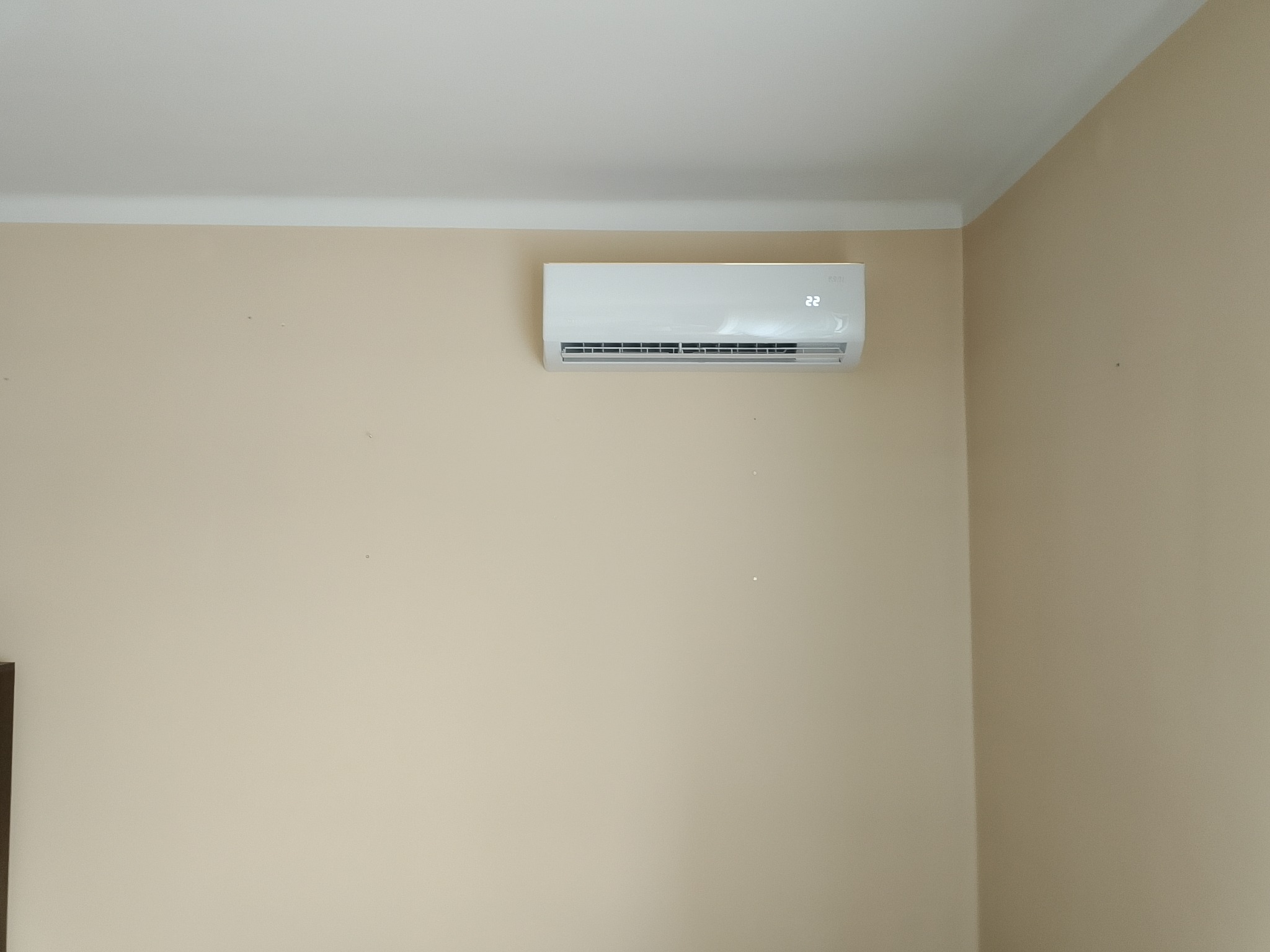 Biały klimatyzator ścienny z wyświetlaczem temperatury 22°C, zamontowany w rogu pokoju na beżowej ścianie pod białym sufitem.