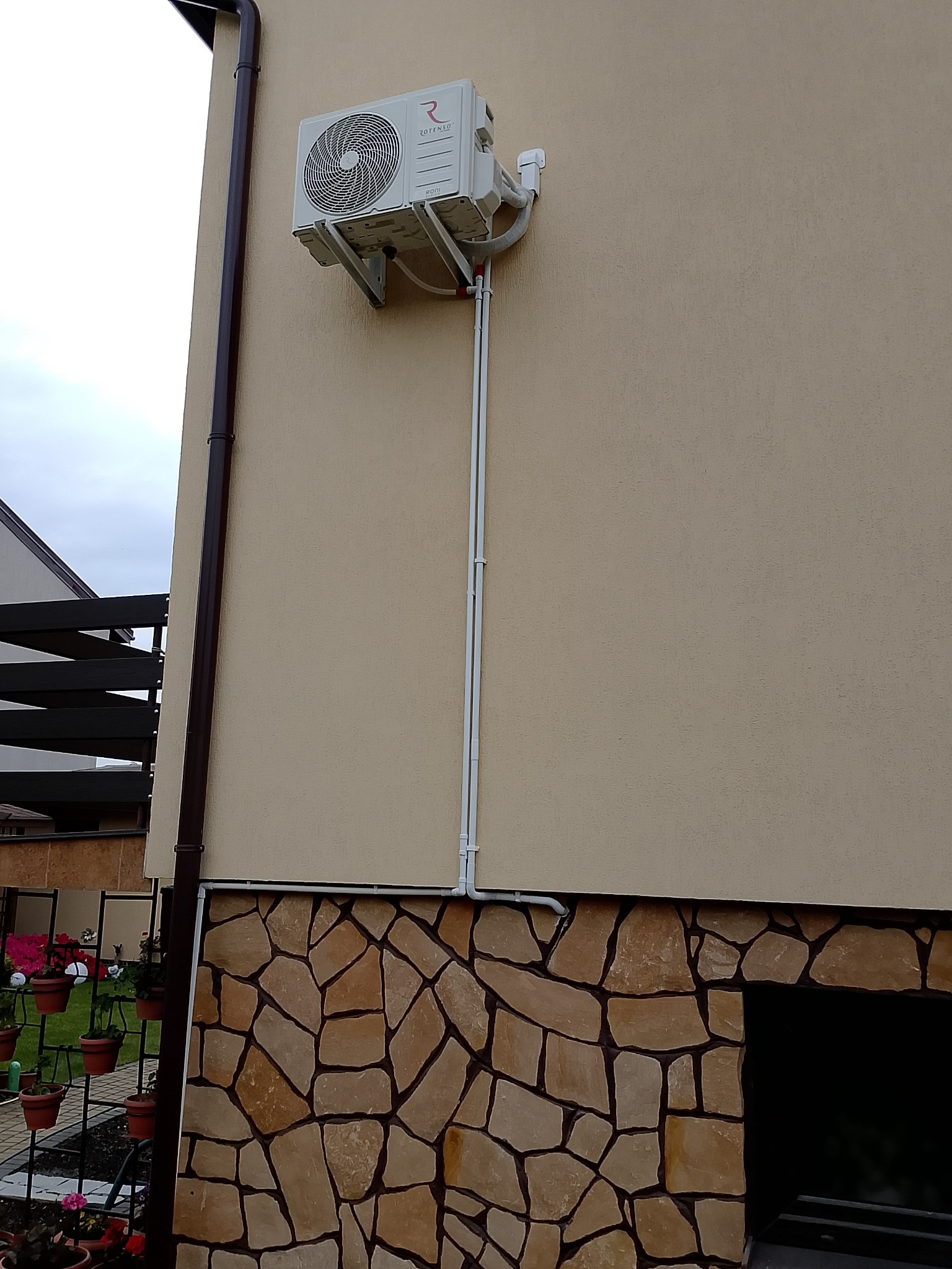 Zamontowany klimatyzator Rotenso na elewacji budynku z kamienną podmurówką. Widoczne rury instalacyjne i rynna. Ujęcie z dołu, lekko z lewej strony.