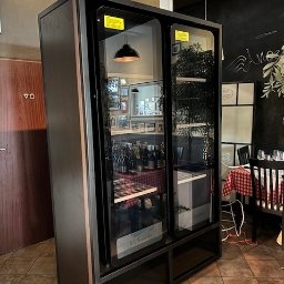 Ślusarstwo-Spawalnictwo Artem Sidorenko - Duża, czarna, przeszklona witryna chłodnicza na wino w restauracji, widoczne butelki wina, stolik z czerwonym obrusem w kratę i drzwi do toalety w tle.