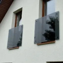 Dwa szklane panele balkonowe zamontowane na elewacji budynku, z widocznym odbiciem drzew w szybach i drewnianymi elementami mocującymi.