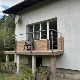 Balkon z balustradą nierdzewną, wykończony panelami imitującymi drewno, z czarnym krzesłem na tle białej ściany budynku i zieleni.