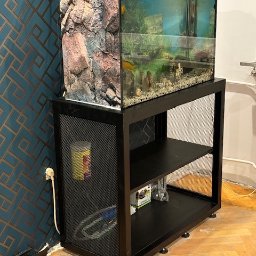 Ślusarstwo-Spawalnictwo Artem Sidorenko - Akwarium z rybkami na metalowym stojaku z siatkowymi bokami i dwiema półkami, widoczne akcesoria na półkach i kabel zasilający w gniazdku.