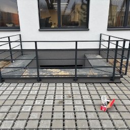 Ślusarstwo-Spawalnictwo Artem Sidorenko - Zabezpieczenie zejścia do piwnicy przy budynku mieszkalnym, wykonane z metalowych profili pomalowanych na czarno, otoczone betonową kostką brukową. Na kostce leży opakowanie po śrubach.