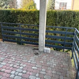 Ciemnoszara balustrada metalowa na tarasie z kostki brukowej w odcieniach czerwieni i szarości, z zielonym żywopłotem w tle.