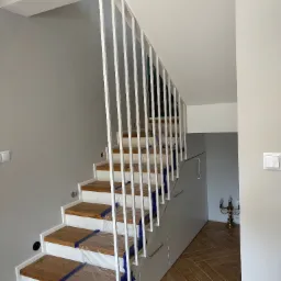 Minimalistyczne schody z drewnianymi stopniami i białą, pionową balustradą metalową. Stopnie zabezpieczone folią. Pod schodami zabudowa meblowa.