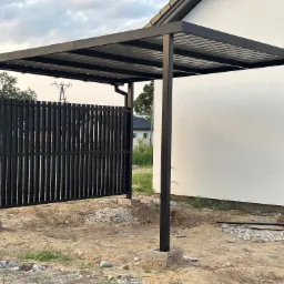 Nowoczesna, czarna konstrukcja zadaszenia z metalu z pionowymi panelami ogrodzeniowymi, osadzona na betonowych fundamentach. Widok na dom w tle.