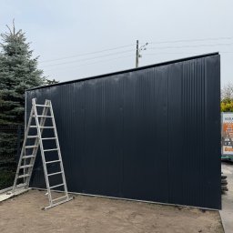 Ślusarstwo-Spawalnictwo Artem Sidorenko - Ciemna, metalowa brama przemysłowa z drabinką opartą o bok, na tle pochmurnego nieba i zieleni. Widoczne pionowe panele z delikatnym żłobieniem.