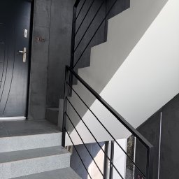 Ślusarstwo-Spawalnictwo Artem Sidorenko - Nowoczesne schody z czarną, metalową balustradą o minimalistycznym designie. Szare płytki schodowe i kontrastujące, białe ściany. Widok z dołu, blisko drzwi wejściowych.