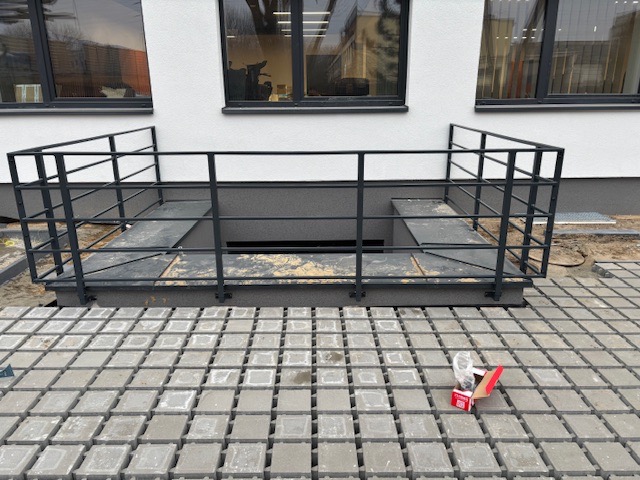 Zabezpieczenie zejścia do piwnicy przy budynku mieszkalnym, wykonane z metalowych profili pomalowanych na czarno, otoczone betonową kostką brukową. Na kostce leży opakowanie po śrubach.