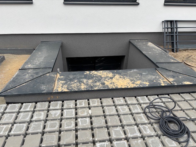 Montaż zejścia do piwnicy z metalową obudową w Łodzi, widoczne ułożone kostki brukowe i przewód elektryczny.