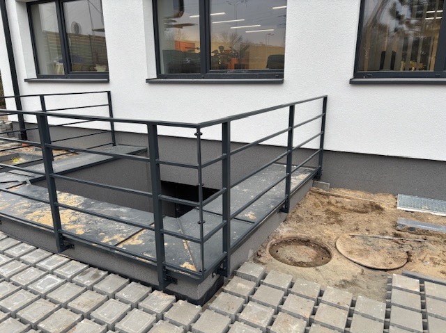 Metalowa konstrukcja zabezpieczająca zejście do piwnicy przy budynku, widoczne niedokończone prace brukarskie i ziemne.