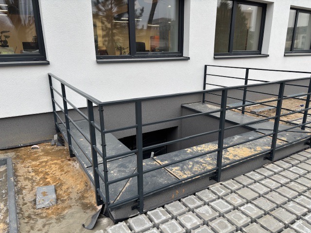 Metalowa barierka zabezpieczająca zejście do piwnicy, widoczna kostka brukowa i fragment elewacji budynku z oknami.
