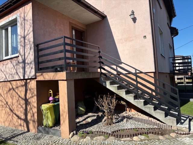 Zewnętrzne schody betonowe z czarną, poziomą balustradą prowadzące do wejścia domu z balkonem, widok z poziomu ogrodu z kostki brukowej.