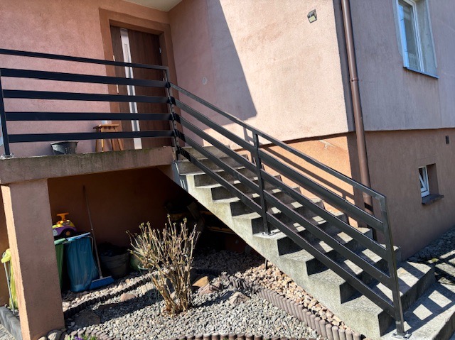 Zewnętrzne schody betonowe z czarną, poziomą balustradą na tle różowej elewacji domu z oknem i drzwiami. U podnóża schodów kamienny ogródek z krzewem.