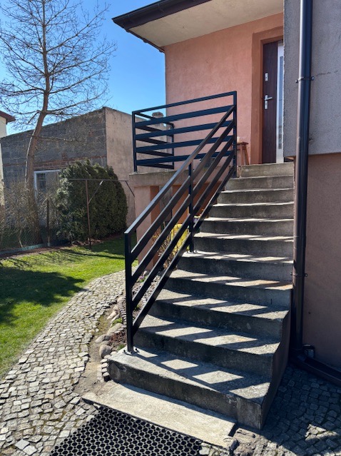 Zewnętrzne schody betonowe z czarną, metalową barierką w Łodzi, prowadzące do drzwi wejściowych budynku. Widoczny trawnik i kostka brukowa.