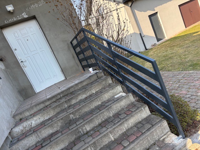 Zewnętrzne schody z szarego betonu z nowoczesną, metalową poręczą w kolorze antracytowym, prowadzące do białych drzwi wejściowych z napisem kredą na ścianie budynku.